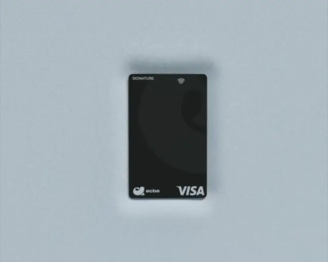 Matte black acba Visa Signature card with a contactless mark, shot top-down on grey linen